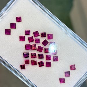 Könnte beinhalten: Eine Sammlung quadratisch geschliffener, tiefroter Edelsteine, präsentiert in einem transparenten Kunststoffbehälter. Die Edelsteine sind auf einer weißen Oberfläche angeordnet, wodurch ihre lebendige Farbe und präzisen Schnitte hervorgehoben werden. Die Edelsteine variieren leicht in der Schattierung.
