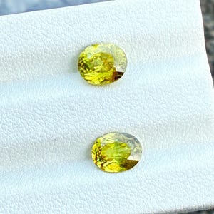 May include: Two round and oval-cut, yellow-green gemstones are displayed on a white textured surface. The gemstones have a brilliant cut, reflecting light. The gems are likely for jewelry making.