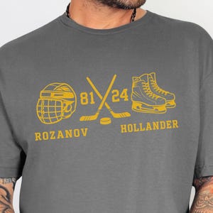 May include: Charcoal gray t-shirt with a gold hockey-themed graphic. The design includes a hockey helmet, crossed hockey sticks, skates, and the numbers "81" and "24". The names "Rozanov" and "Hollander" are also featured.