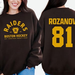 May include: Dark chocolate sweatshirt with "RAIDERS BOSTON HOCKEY" text in gold. The front features a logo and the text "EST. NINETEEN TWENTY-FOUR". The back displays "ROZANOV 81" in gold.