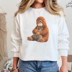 Puede incluir: Sudadera blanca con una ilustraci&oacute;n detallada de un orangut&aacute;n abrazando a un beb&eacute; mono. El orangut&aacute;n tiene pelaje marr&oacute;n y el beb&eacute; mono es gris. El texto "White Sweatshirt" est&aacute; en la esquina superior derecha.