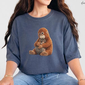 Puede incluir: Camiseta azul denim con un gr&aacute;fico de una madre orangut&aacute;n abrazando a su beb&eacute;. Los orangutanes est&aacute;n representados en tonos marrones y beige. La palabra "denim" est&aacute; escrita en cursiva en la esquina superior derecha.