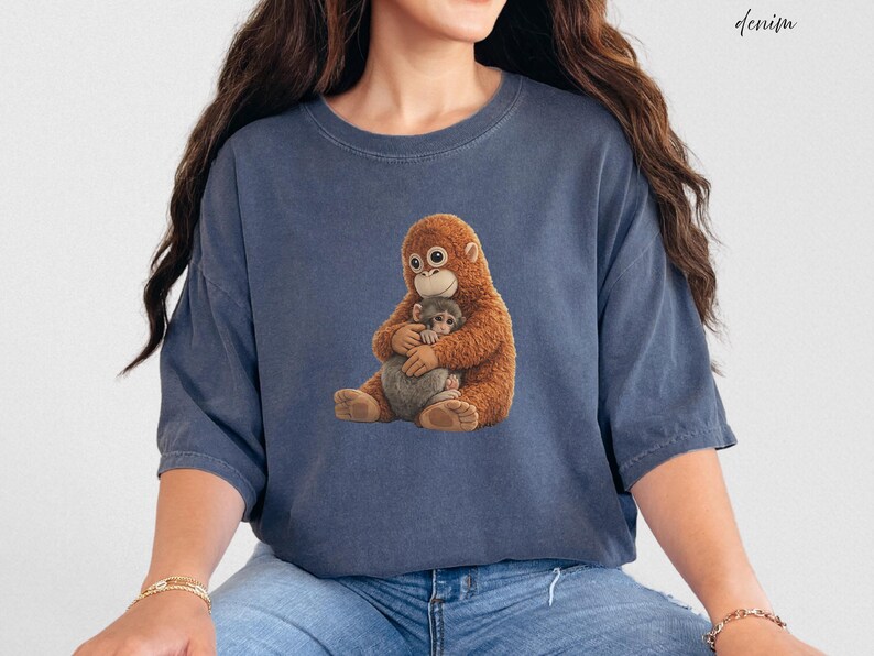 Puede incluir: Camiseta azul denim con un gr&aacute;fico de una madre orangut&aacute;n abrazando a su beb&eacute;. Los orangutanes est&aacute;n representados en tonos marrones y beige. La palabra "denim" est&aacute; escrita en cursiva en la esquina superior derecha.