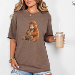 Puede incluir: Camiseta marr&oacute;n con un dibujo de un orangut&aacute;n abrazando a un beb&eacute; orangut&aacute;n. La camiseta es de color marr&oacute;n apagado. La palabra "Espresso" est&aacute; escrita en cursiva en la esquina superior derecha. Un vaso transparente con pajita se sostiene en la mano derecha.