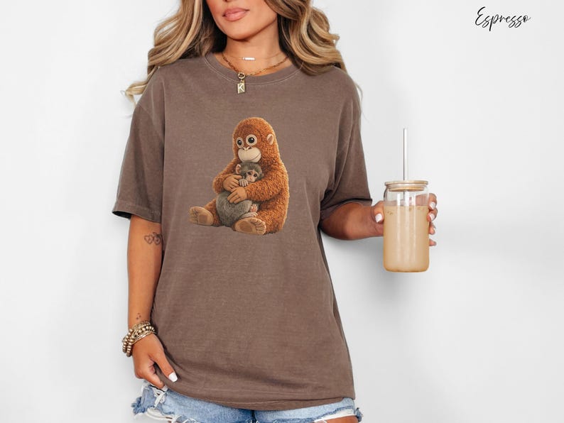 Puede incluir: Camiseta marr&oacute;n con un dibujo de un orangut&aacute;n abrazando a un beb&eacute; orangut&aacute;n. La camiseta es de color marr&oacute;n apagado. La palabra "Espresso" est&aacute; escrita en cursiva en la esquina superior derecha. Un vaso transparente con pajita se sostiene en la mano derecha.