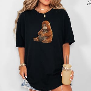 Puede incluir: Camiseta negra con un gr&aacute;fico de un orangut&aacute;n marr&oacute;n abrazando a un animal gris. La palabra "black" est&aacute; escrita en cursiva en la esquina superior derecha. La camiseta es de estilo casual y oversize.