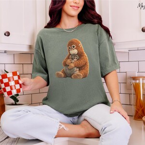 Puede incluir: Una camiseta verde salvia con un gr&aacute;fico de un orangut&aacute;n marr&oacute;n abrazando a un beb&eacute; orangut&aacute;n gris. La camiseta est&aacute; hecha de un material suave. La persona sostiene una taza a cuadros rojos y blancos.