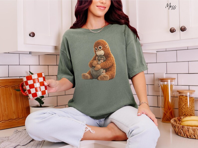 Puede incluir: Una camiseta verde salvia con un gr&aacute;fico de un orangut&aacute;n marr&oacute;n abrazando a un beb&eacute; orangut&aacute;n gris. La camiseta est&aacute; hecha de un material suave. La persona sostiene una taza a cuadros rojos y blancos.
