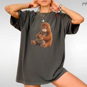 Puede incluir: Camiseta gris oscuro con un gr&aacute;fico de una madre orangut&aacute;n abrazando a su beb&eacute;. Los orangutanes est&aacute;n representados en tonos marrones y beige. La camiseta tiene un corte holgado y mangas cortas.