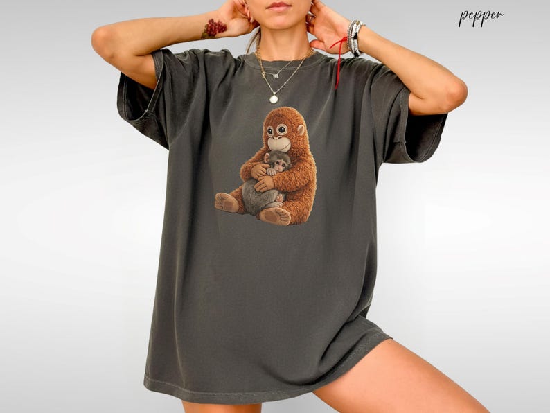 Puede incluir: Camiseta gris oscuro con un gr&aacute;fico de una madre orangut&aacute;n abrazando a su beb&eacute;. Los orangutanes est&aacute;n representados en tonos marrones y beige. La camiseta tiene un corte holgado y mangas cortas.