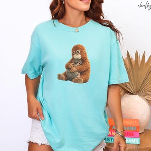 Puede incluir: Camiseta azul claro con un gr&aacute;fico de un orangut&aacute;n marr&oacute;n abrazando a un animal gris. El texto "chalky mint" est&aacute; escrito en cursiva. La camiseta es de estilo casual y relajado.