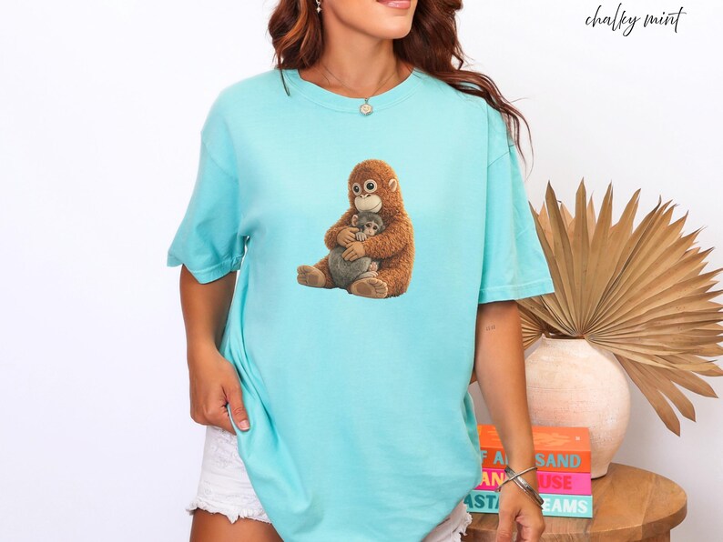 Puede incluir: Camiseta azul claro con un gr&aacute;fico de un orangut&aacute;n marr&oacute;n abrazando a un animal gris. El texto "chalky mint" est&aacute; escrito en cursiva. La camiseta es de estilo casual y relajado.