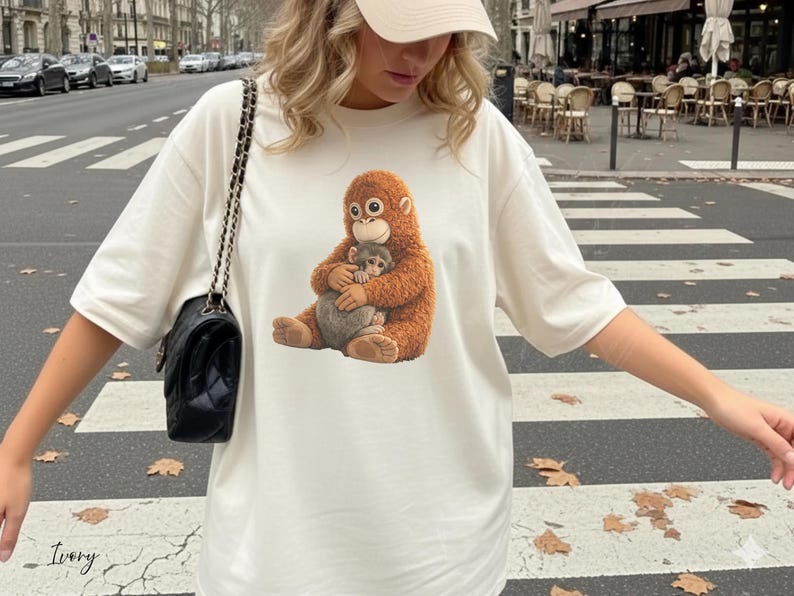 Puede incluir: Camiseta de color marfil con una ilustraci&oacute;n detallada de un orangut&aacute;n marr&oacute;n abrazando a un mono m&aacute;s peque&ntilde;o. El dise&ntilde;o est&aacute; centrado en la camiseta. La palabra "Ivory" est&aacute; escrita en cursiva en la parte inferior izquierda de la camiseta.