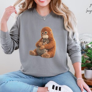 Puede incluir: Una sudadera gris de manga larga con un orangut&aacute;n de dibujos animados y un animal m&aacute;s peque&ntilde;o. El orangut&aacute;n es marr&oacute;n y peludo, y el animal m&aacute;s peque&ntilde;o es gris. El texto "grey long sleeve" est&aacute; escrito en cursiva en la esquina superior derecha.