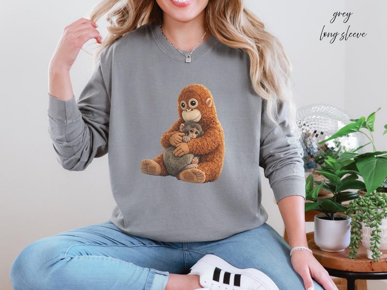 Puede incluir: Una sudadera gris de manga larga con un orangut&aacute;n de dibujos animados y un animal m&aacute;s peque&ntilde;o. El orangut&aacute;n es marr&oacute;n y peludo, y el animal m&aacute;s peque&ntilde;o es gris. El texto "grey long sleeve" est&aacute; escrito en cursiva en la esquina superior derecha.