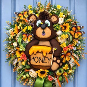 May include: A whimsical wreath featuring a brown bear holding a honey pot with the word "HONEY". The wreath is adorned with sunflowers, butterflies, and various flowers in shades of yellow, orange, and green. Decorative ribbons add to the festive design.