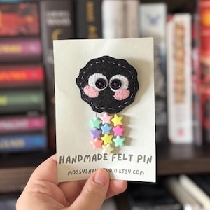 Puede incluir: Un pin de fieltro hecho a mano con un diseño de duende de hollín negro con mejillas rosas y ojos morados, sujeto a una tarjeta. El pin tiene un grupo de adornos en forma de estrella coloridas. La tarjeta dice "HANDMADE FELT PIN" y el nombre de la tienda Etsy.