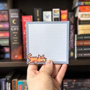 May include: A square, light blue sticky note with a grid pattern and a pixelated orange cat graphic in the corner. The sticky note is held in front of a bookshelf filled with books.