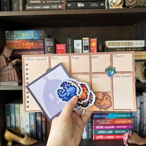 May include: A beige weekly planner with pixelated graphics and the words "Main Quest" and "Side Quest." The planner has boxes for each day of the week. A hand holds pixelated stickers of a blue gem, a red gem, and a chicken.