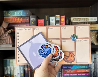 May include: A beige weekly planner with pixelated graphics and the words "Main Quest" and "Side Quest." The planner has boxes for each day of the week. A hand holds pixelated stickers of a blue gem, a red gem, and a chicken.