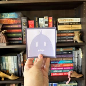May include: A square sticker featuring a pixelated ghost design in shades of purple and white. The sticker is held in front of a bookshelf filled with various books. The ghost has a simple, minimalist design.
