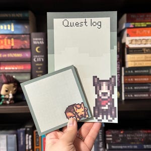 May include: A set of pixel art-style notepads with a light green background. One notepad reads "Quest log" in pixelated text. The other notepad features a pixelated dog and a person. The notepads are held in a person's hand.