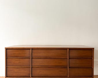 Puede incluir: Una cómoda moderna de mediados de siglo con nueve cajones. La cómoda de madera es de color marrón cálido y tiene forma rectangular. La cómoda tiene patas cónicas y un diseño limpio y minimalista, ideal para el almacenamiento en el dormitorio.