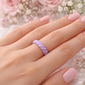 May include: A lilac silicone ring with a twisted rope design. The ring is on a finger, with a pale pink manicure. The background features pink roses and white flowers.