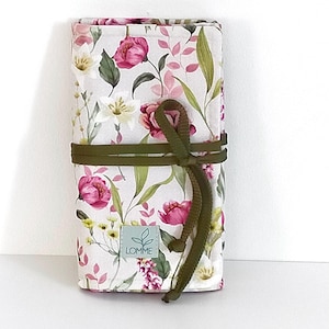 May include: A floral print fabric roll featuring pink peonies, green leaves, and yellow wildflowers on a white background. The roll is secured with a dark green cord. A small label with the text "LOMME" is visible.