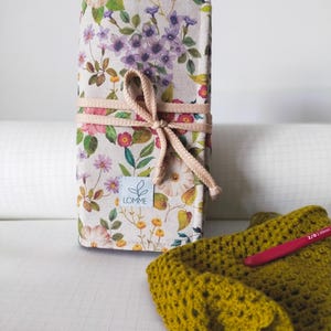 Puede incluir: Un estuche para ganchillos con estampado floral, atado con una cinta rosa. El estuche presenta un patrón de flores moradas, rosas y amarillas sobre un fondo crema. Un artículo de ganchillo verde amarillento y un ganchillo rojo están junto al estuche.