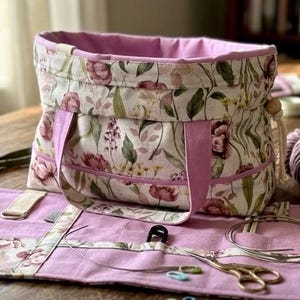 May include: A floral print knitting project bag with matching accessories, including a needle roll, scissors, and yarn. The bag has a pink interior and handles. A skein of yarn and a mug are also visible.