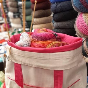 Op de afbeelding: Een beige en roze garentas gevuld met kleurrijke garenbollen en houten breinaalden. De tas heeft roze randen en handvatten, met een verscheidenheid aan garenkleuren zichtbaar, waaronder oranje, roze en blauw. Stapels garen zijn op de achtergrond.