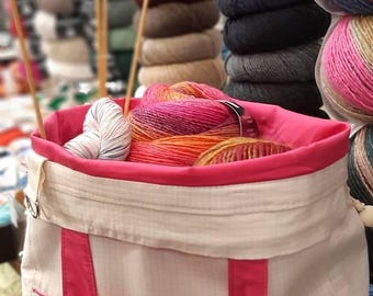 May include: A beige and pink yarn tote bag filled with colorful yarn skeins and wooden knitting needles. The bag has pink trim and handles, with a variety of yarn colors visible, including orange, pink, and blue. Stacks of yarn are in the background.