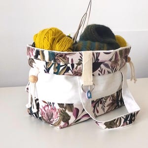 Op de afbeelding: Een stoffen opbergmand met een bloemenprint in roze, groene en donkerbruine botanische motieven op een crèmekleurige achtergrond. De mand heeft een witte middenband, witte katoenen hengsels, houten kralen en bevat diverse bollen wol.