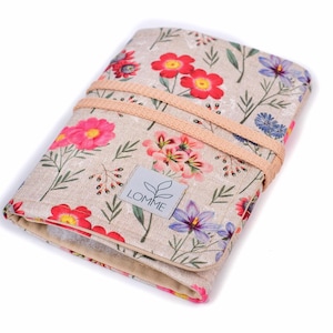 Puede incluir: Un organizador de tela rectangular con base beige y un estampado floral colorido de flores rojas, rosas y moradas con tallos verdes. El artículo está sujeto por dos bandas elásticas de color melocotón claro y tiene una etiqueta con LOMME.