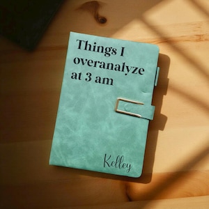 May include: A teal journal with the text "Things I overanalyse at 3 am" and the name "Kelley" printed on the cover. The journal has a gold clasp and is on a wooden surface.