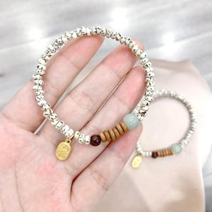 May include: Two beaded bracelets. The main bracelet has white beads with black speckles, wooden beads, a small gold charm, and a light green bead. The second bracelet is similar. The bracelets are on a light pink surface.