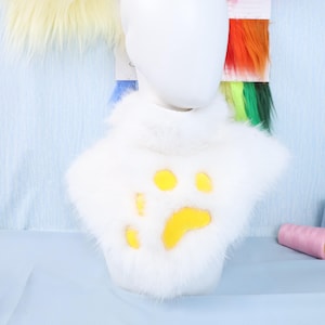 May include: A white faux fur neck piece with a yellow paw print design. The neck piece is displayed on a white mannequin head. Colorful fabric swatches are visible in the background.