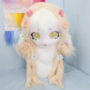May include: A white and tan furry costume head with large, expressive eyes and floral accents. The head has long, flowing hair and is decorated with small, colorful flowers. The eyes are yellow with a cross design. Sample hair swatches are visible in the background.