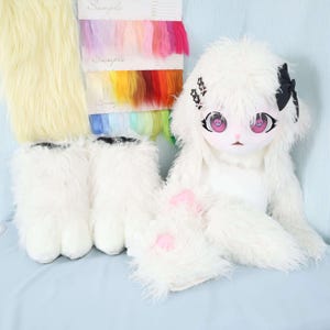May include: A white furry doll with a matching head covering, paws, and feet. The doll has large eyes with pink irises and black accents. A swatch of colorful fur samples and a swatch of yellow fur are also present.