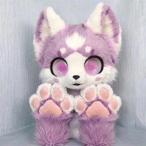 May include: A plush toy resembling a fox or wolf with long, soft lavender and white fur. It features large, expressive eyes with heart-shaped pupils, pointed ears, and two raised paws with light pink pads. The face has a white muzzle and forehead.