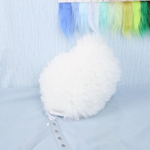 May include: A fluffy, white, heart-shaped object with a clear strap featuring snap closures. The background includes a color swatch of various shades of blue, green, and yellow. The object appears to be a decorative item or accessory.