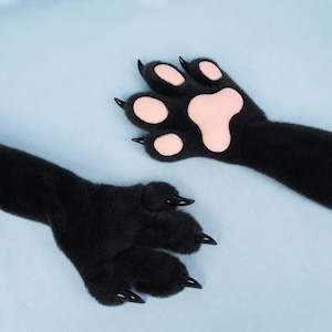 May include: A pair of black plush animal paw gloves featuring soft black faux fur, light pink padded paw pads, and sharp, glossy black claws extending from each of the four fingers. The design mimics a feline or predatory animal paw.