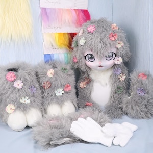 May include: A doll with a gray fur body and head, adorned with small, colorful flowers. The doll has large, expressive eyes and is surrounded by matching fur accessories, including gloves and leg warmers. Samples of colorful fur are visible in the background.