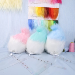 May include: Three fluffy, two-toned faux fur tails in pink, mint green, and blue, each with a white base. Clear adjustable straps are attached. A color swatch card with the word "Sample" is visible, along with spools of thread.