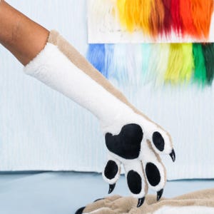 Könnte beinhalten: Ein langer Handschuh aus weichem Kunstfell in Form einer Tierpfote. Der Handschuh hat eine weiße Unterseite, eine hellbraune Oberseite, schwarze Pfotenballen und spitze, schwarze Krallen an den Fingerspitzen. Das Material ist weich und strukturiert.