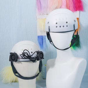 May include: Two white helmets with black straps and vents. One helmet has two black fans attached, connected by wires. The helmets are displayed on mannequin heads. A colorful swatch of hair samples is in the background.