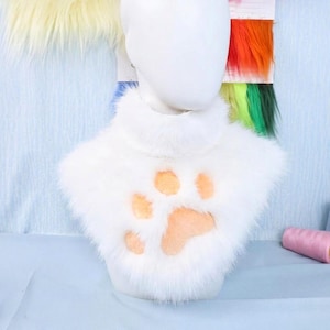 May include: A white faux fur neck warmer featuring a high collar and a large, light peach-colored paw print design centered on the front. The soft, plush material covers the neck and upper chest area, providing a textured, fuzzy appearance.