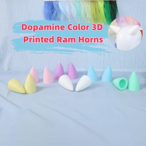 May include: A collection of 3D-printed ram horns in various pastel colors, including yellow, pink, purple, blue, white, and green. The text "Dopamine Color 3D Printed Ram Horns" is displayed above the horns. The horns are arranged on a light blue surface.