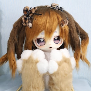 May include: A doll with a large head, big eyes, and a furry appearance. The doll has brown and orange hair styled in pigtails, adorned with small accessories. It wears furry paw-like gloves and has a light-colored face with detailed features.